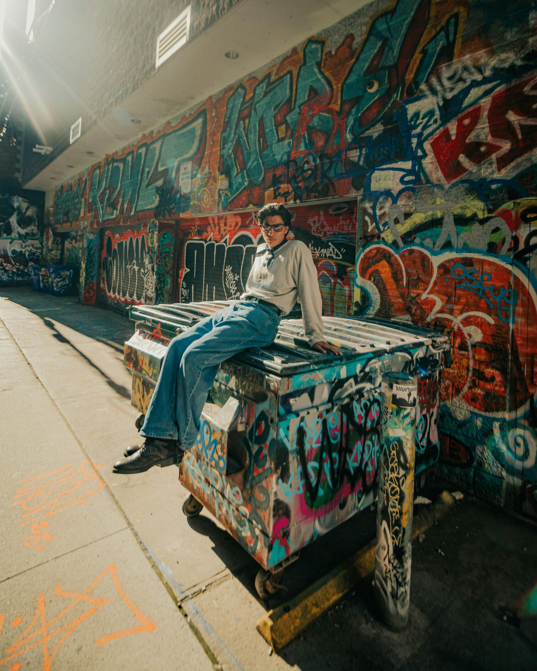 A young man wearing eyeglasses sits on a graffiti-covered dumpster in a vibrant urban alley.