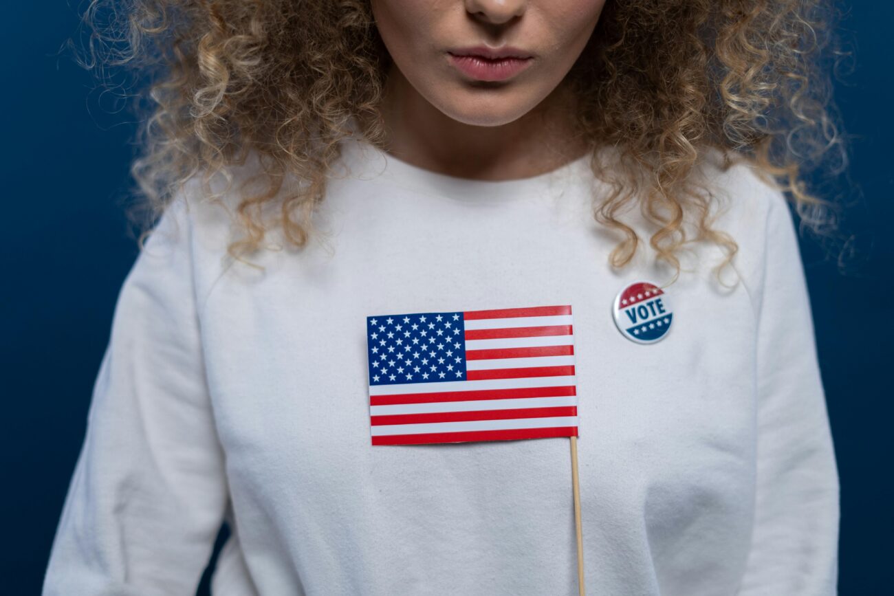 A woman with curly hair holding an American flag and wearing a vote pin against a blue background.