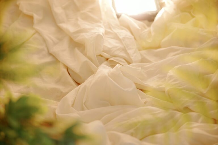 Soft, cream-colored silk bedding softly illuminated by warm light, creating a cozy and luxurious atmosphere with gentle fabric folds and blurred greenery in the foreground.