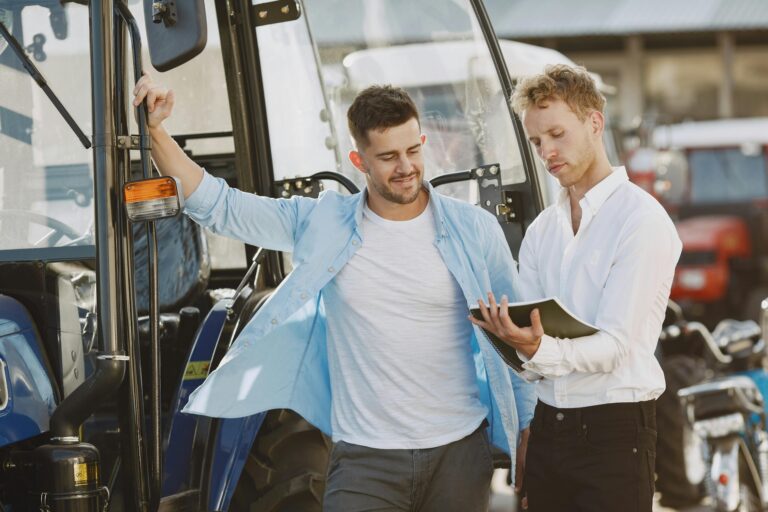 Two businessmen discussing a vehicle purchase outdoors with a tractor in the background.